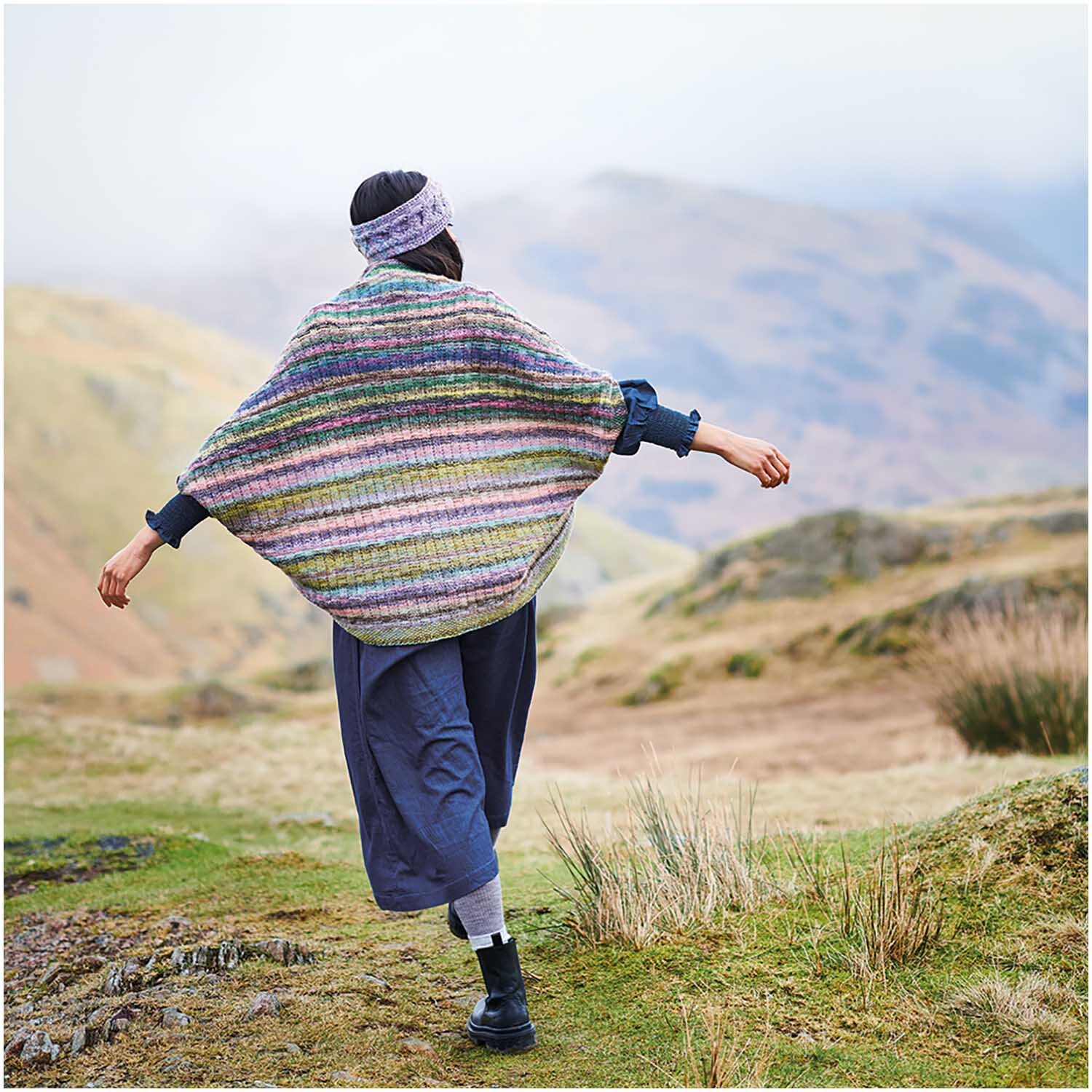Strickset Poncho Modell 18: Person in buntem Poncho in bergiger Landschaft, von hinten gesehen.
