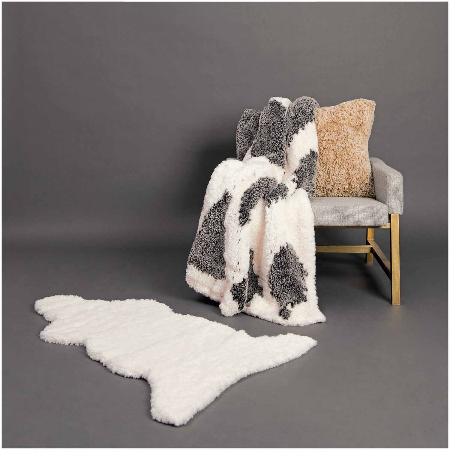 Creative Petit Lapin Super Chunky Decke in Taupe-Weiß auf einem grauen Sessel, daneben ein passender Teppich. Ideal für gemütliche Wohnräume.