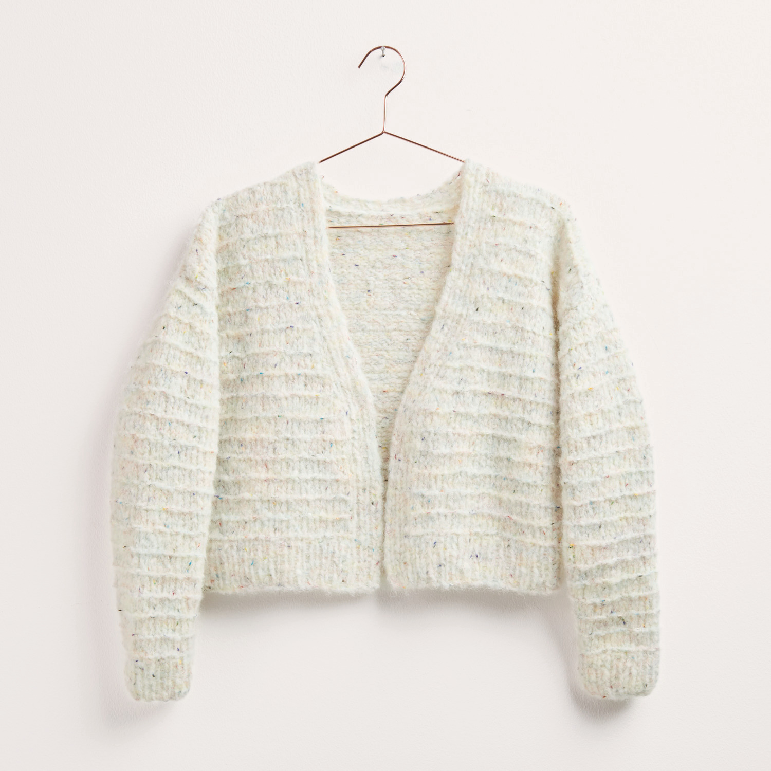 Strickset Jacke Modell 01: Cremefarbene Strickjacke aus weichem Garn, präsentiert auf einem Kleiderbügel. Ideal für Herbst-Winter. Marke: Rico Design.