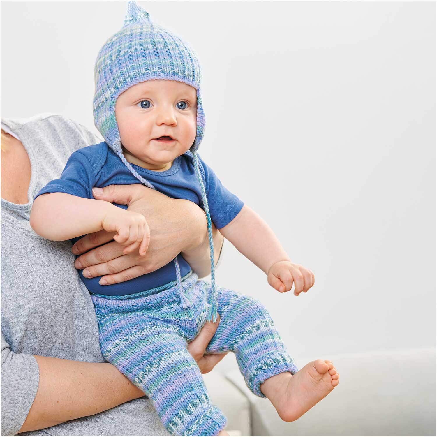 Rico Baby Dream dk A Luxury Touch: Ein Baby in gestrickter Kleidung in Blau- und Grüntönen, gehalten von einer Person.