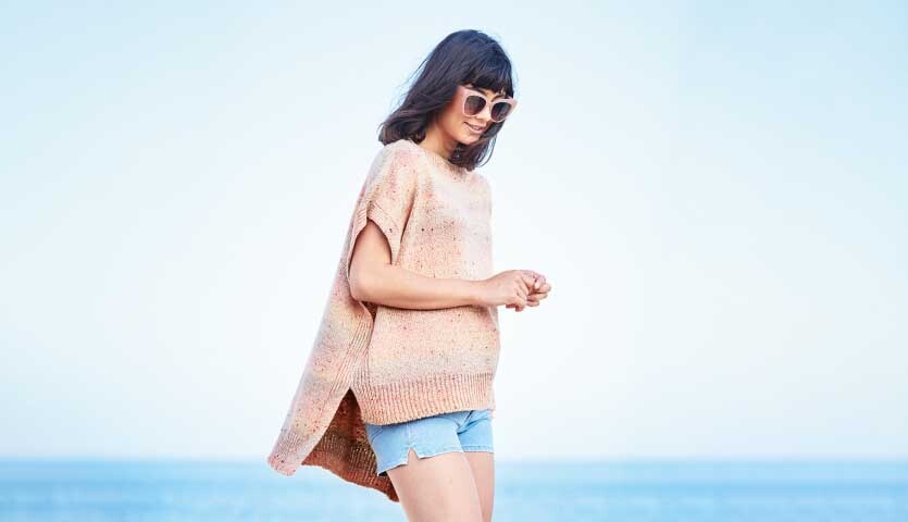Strickanleitung Shirt aus Fashion Cotton Light & Long Tweed dk: Frau am Strand in lockerem, hellen Strickshirt und Shorts, mit Sonnenbrille.
