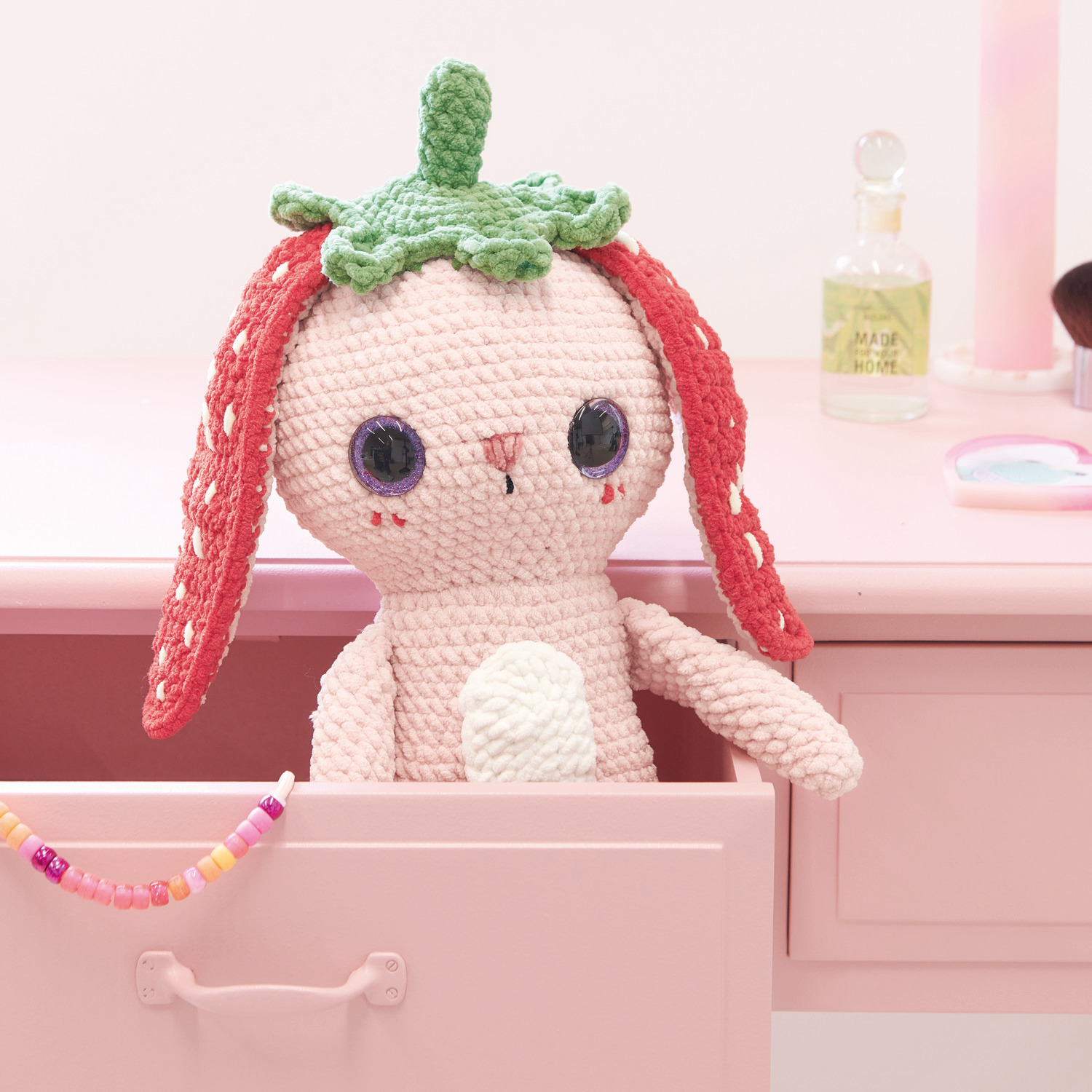 Häkelset Strawberry Bunny Modell 03: Ein gehäkelter Hase mit Erdbeerhut sitzt in einer rosa Schublade.