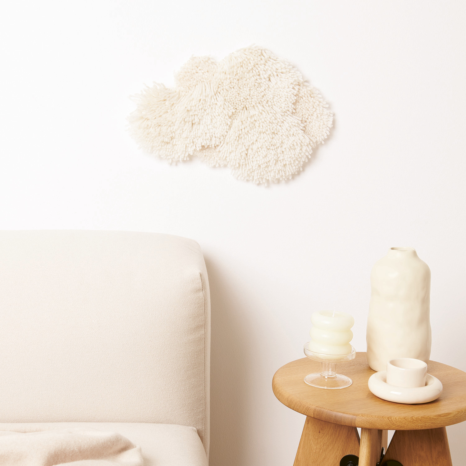 Rico Hooki Hooki Knüpfgarn, offwhite, dekoriert eine Wand über einem Sofa. Daneben ein Tisch mit Vase und Kerzen.