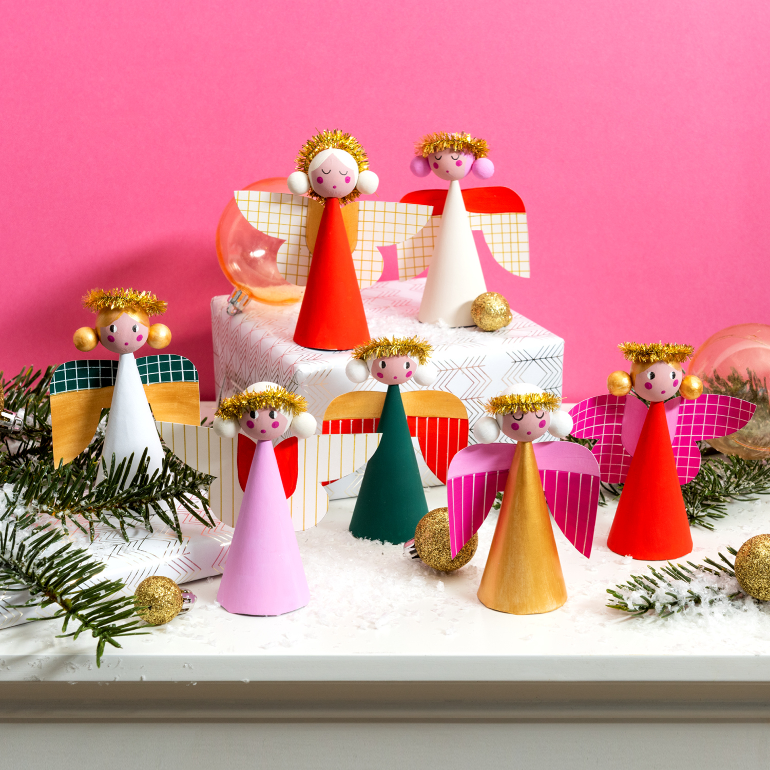 Bastelanleitung Engelfiguren: Bunte Engel aus Papier mit goldenen Kronen vor rosa Hintergrund, umgeben von Tannenzweigen und Weihnachtskugeln.
