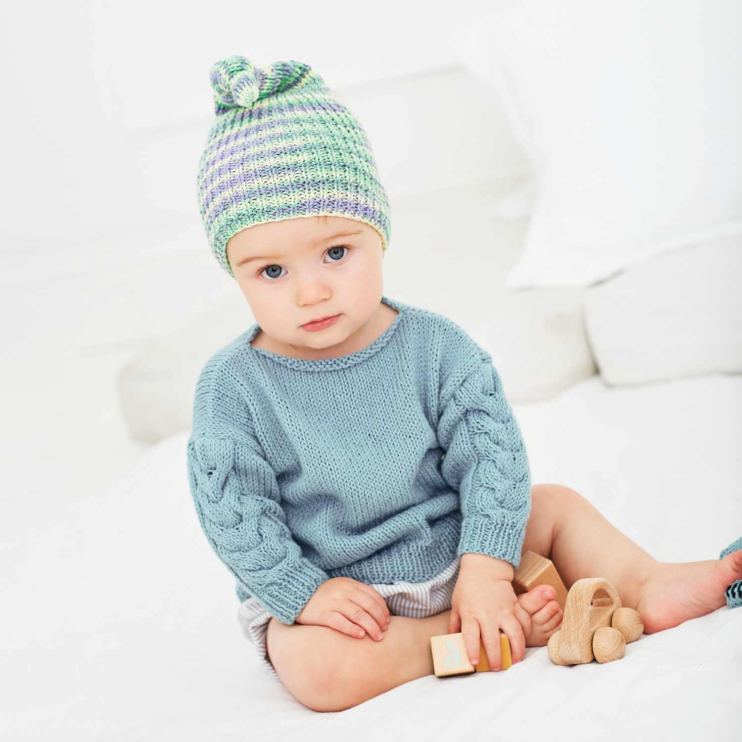 Strickanleitung Mütze/Pullover aus Baby Cotton Soft: Kind in gestricktem Pullover und Mütze, spielt mit Holzspielzeug auf weißem Hintergrund.