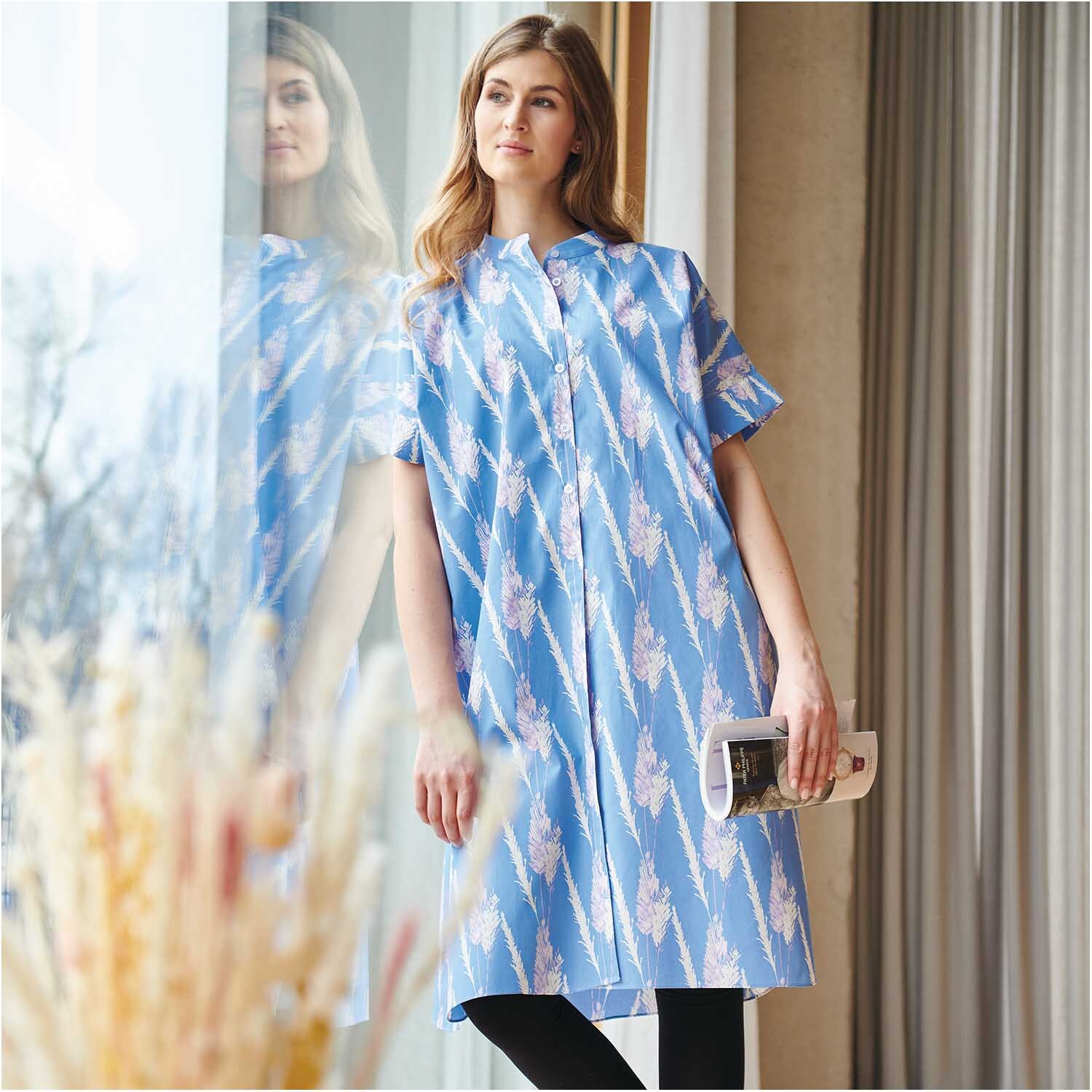 Stoffabschnitt Druckstoff Pampasgras azur zeigt eine Frau in einem blauen Kleid mit Pampasgras-Muster, stehend vor einem Fenster.