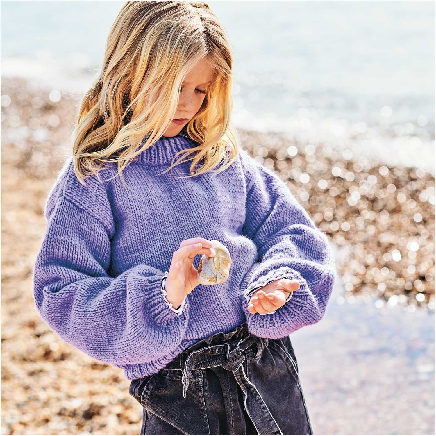 Essentials Super aran: Kind am Strand in lila Pullover von Rico Design, hält Muschel in der Hand.
