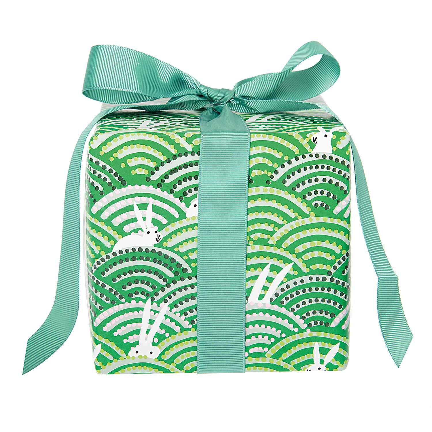 Paper Poetry Geschenkpapier zeigt Hasen im Feld mit grünem Muster, verpackt mit Schleife. Ideal für Ostern. Marke: Rico Design.