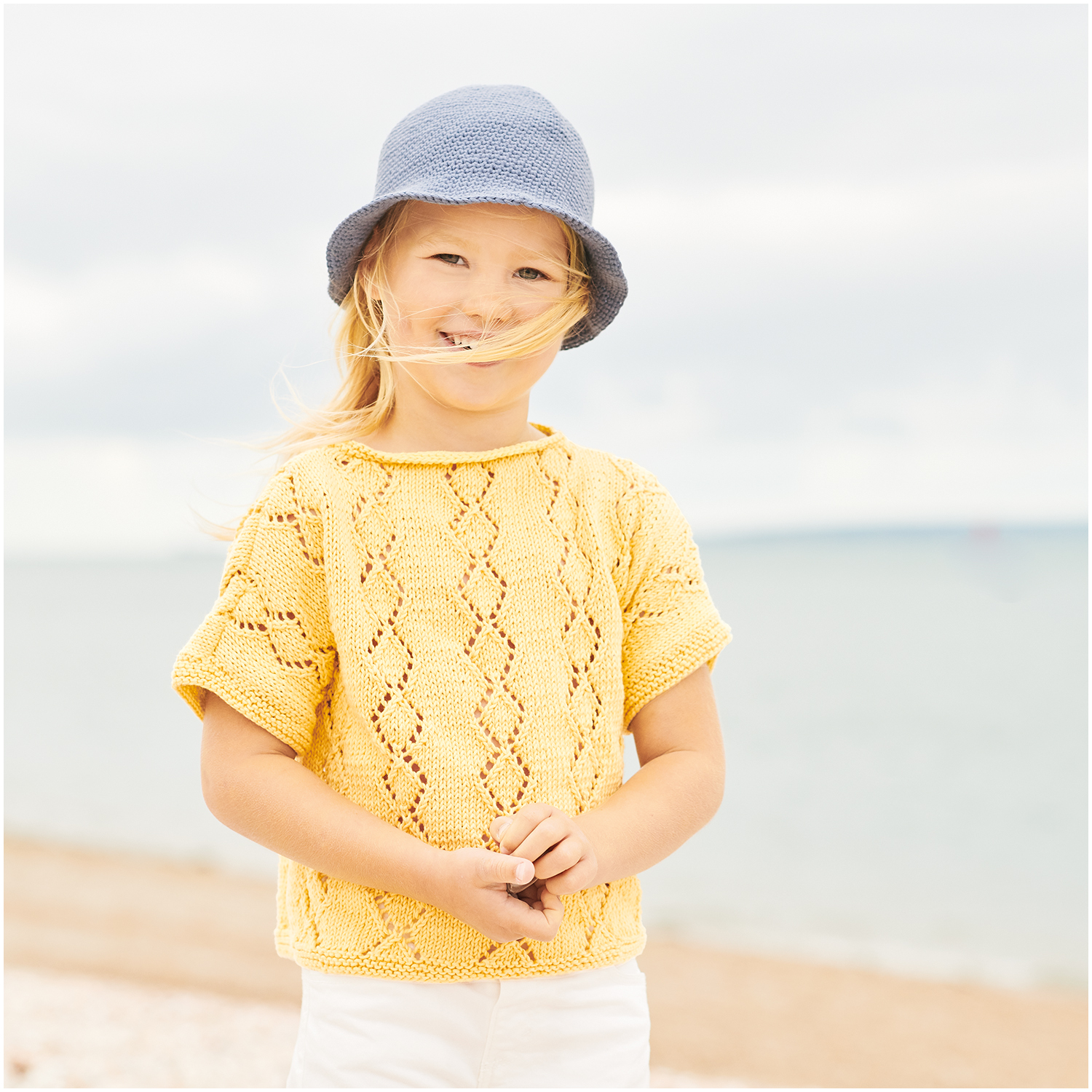Essentials Organic Cotton dk: Kind am Strand in gestricktem, senffarbenem Oberteil und blauem Hut. Ideal für sommerliche Tage.