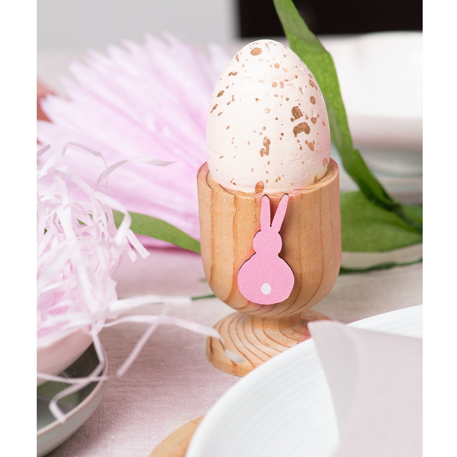 DIY Set Osterdeko bunt: Ein Ei im Holzhalter mit rosa Hasenmotiv, umgeben von Ostergras auf einem Tisch.