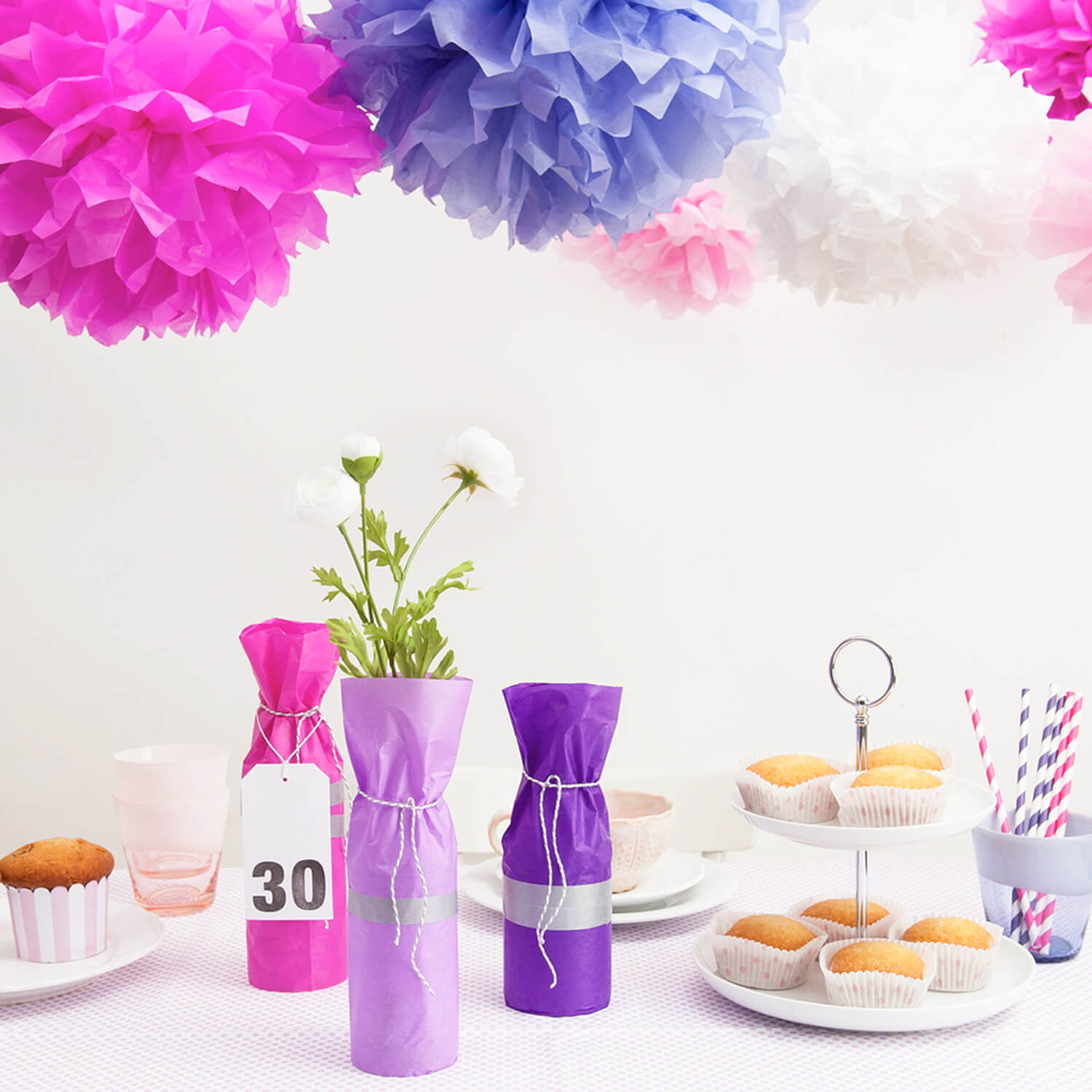 Bastelanleitung Partydeko selber machen: Bunte Pompons und dekorierte Flaschen auf einem Tisch mit Cupcakes und Blumen.