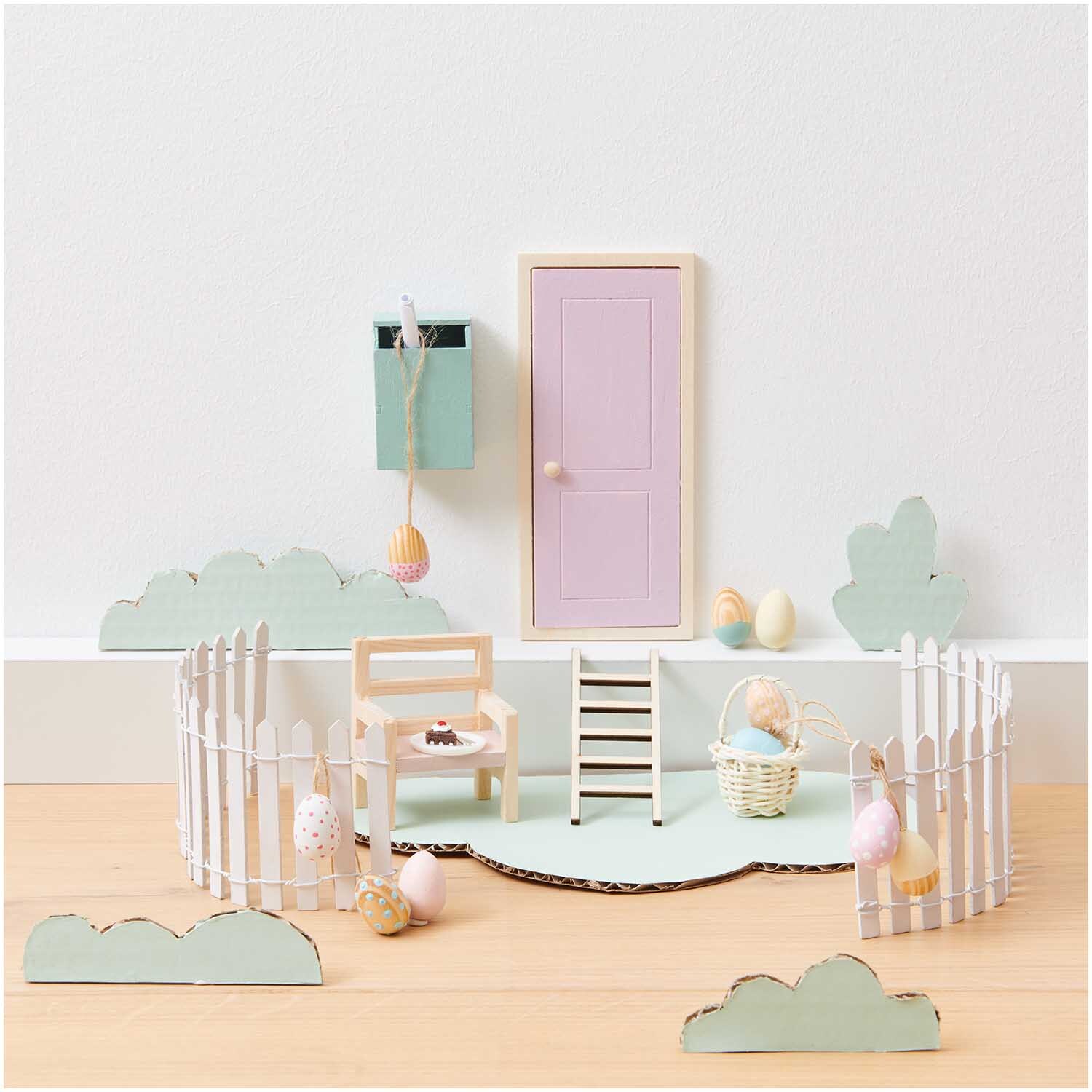 Miniatur Korb mit Henkel in einer pastellfarbenen Szene mit Zäunen, Tür und Ostereiern.