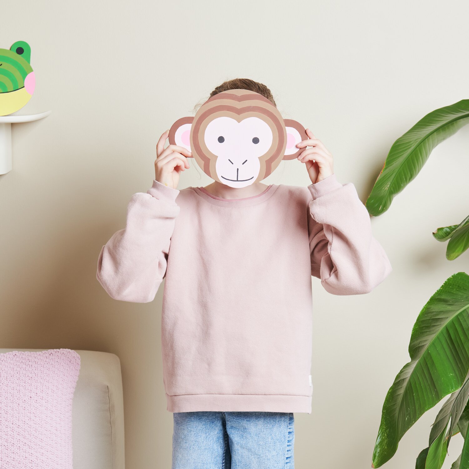 Malen nach Zahlen Affe: Kind in rosa Pullover hält Affenmaske vor Gesicht, neben grüner Pflanze.