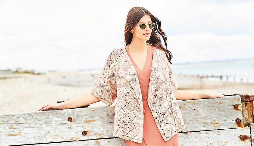 Strickanleitung Jacke aus Fashion Cotton Double Printed dk: Frau am Strand in eleganter, gemusterter Jacke, die über einem korallfarbenen Kleid getragen wird.