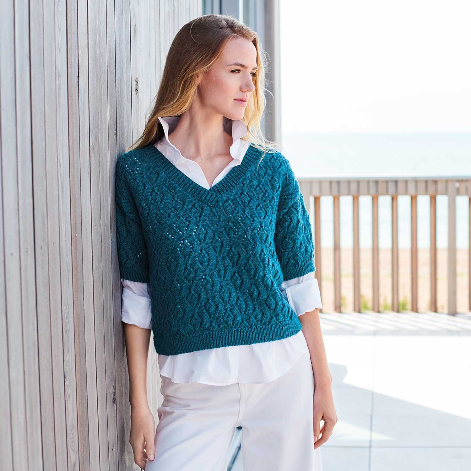 Strickanleitung Shirt aus Creative Silky Touch dk: Frau trägt blaues Strickshirt über weißer Bluse, lehnt an Holzwand.