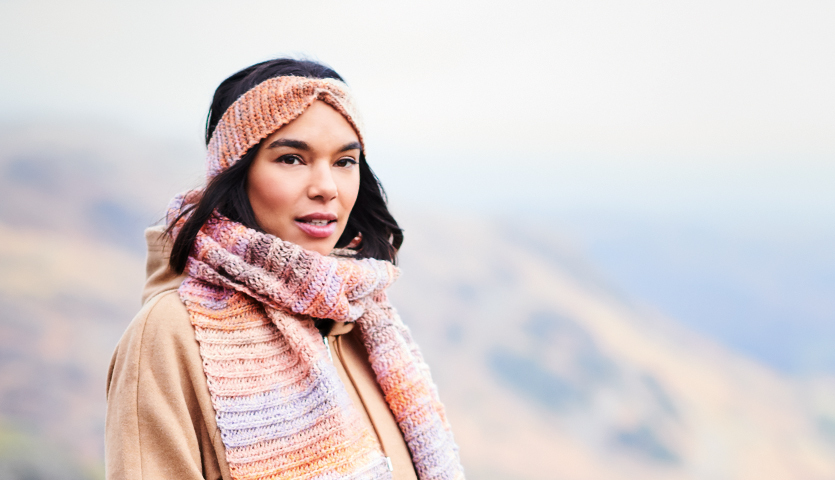 Häkelanleitung Stirnband & Schal aus Creative Chic-Unique dk: Frau trägt gestricktes Stirnband und Schal in Pastelltönen vor nebliger Berglandschaft.