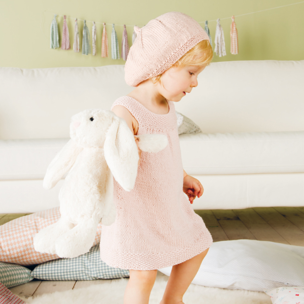 Strickanleitung Rico Baby 19 zeigt ein Kind in rosa Kleid und Mütze, das ein Plüschtier hält.