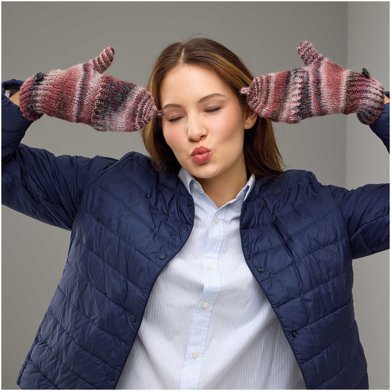 Strickset Fäustlinge Modell 27: Frau in blauer Jacke trägt gestrickte, bunte Fäustlinge und posiert fröhlich.