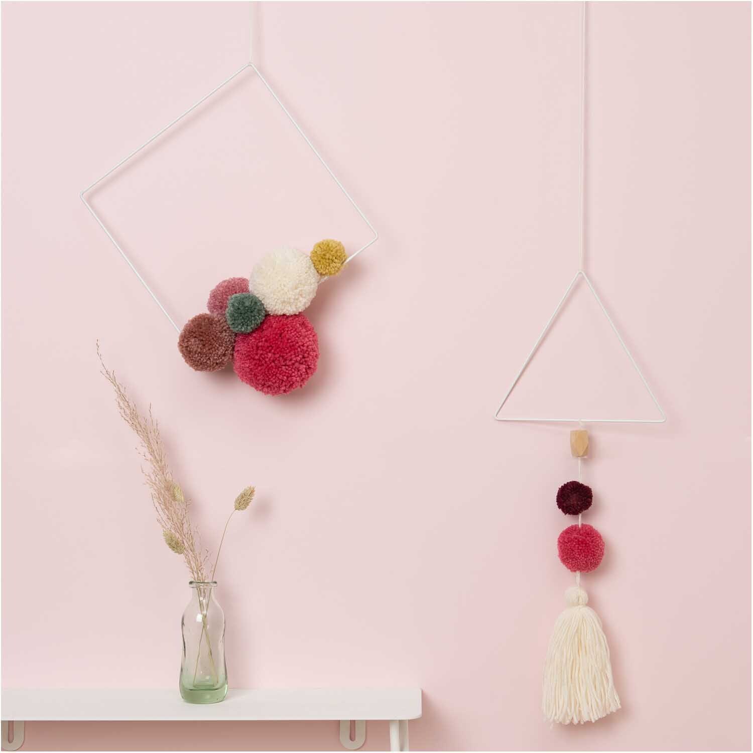 Metallring Quadrat weiß von Rico Design mit bunten Pompons, dekorativ an einer rosa Wand hängend.