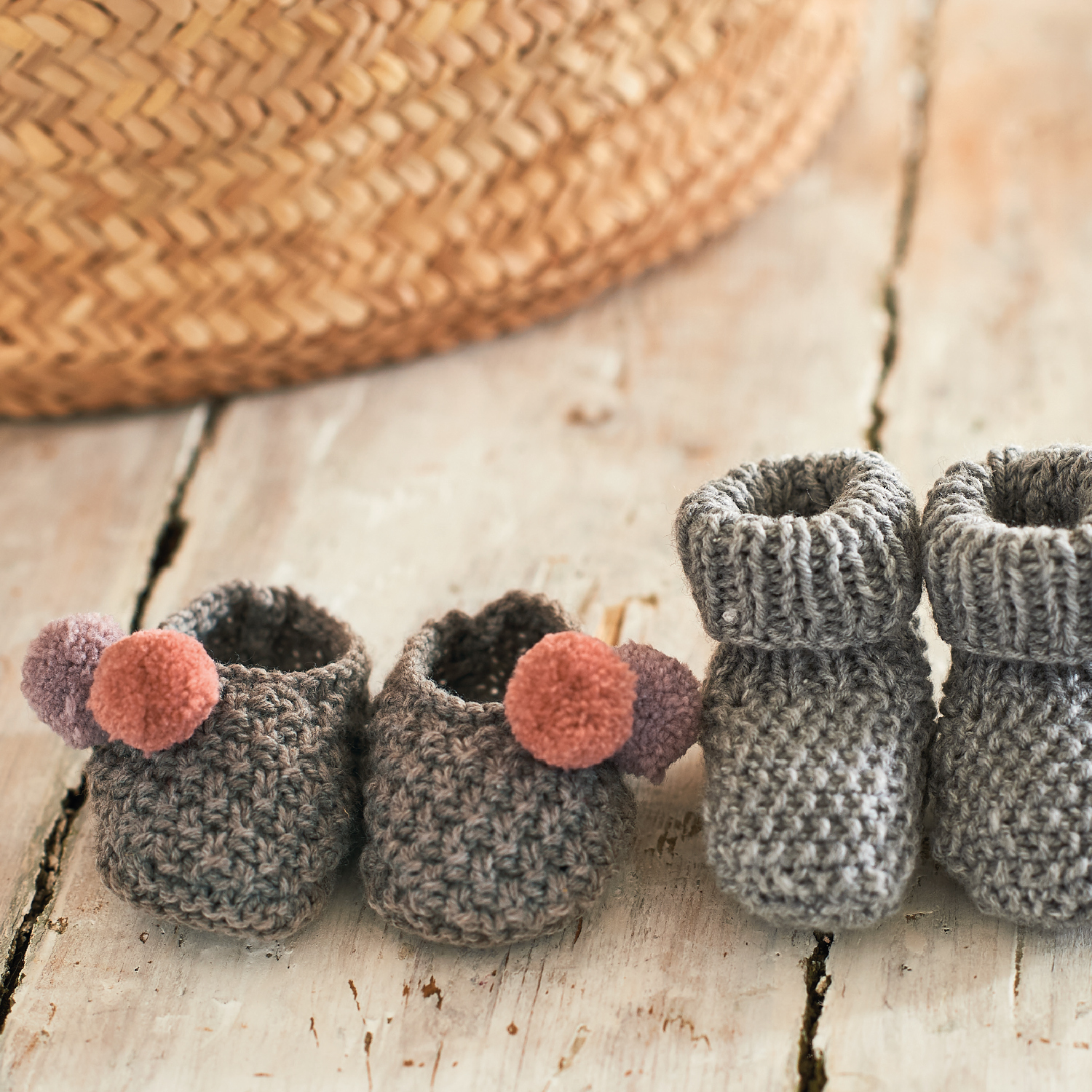 Strickanleitung Babysocken aus Rico Baby Classic dk: Zwei Paar graue Babysocken auf Holztisch, eines mit rosa Pompons, ideal für warme Babyfüße.