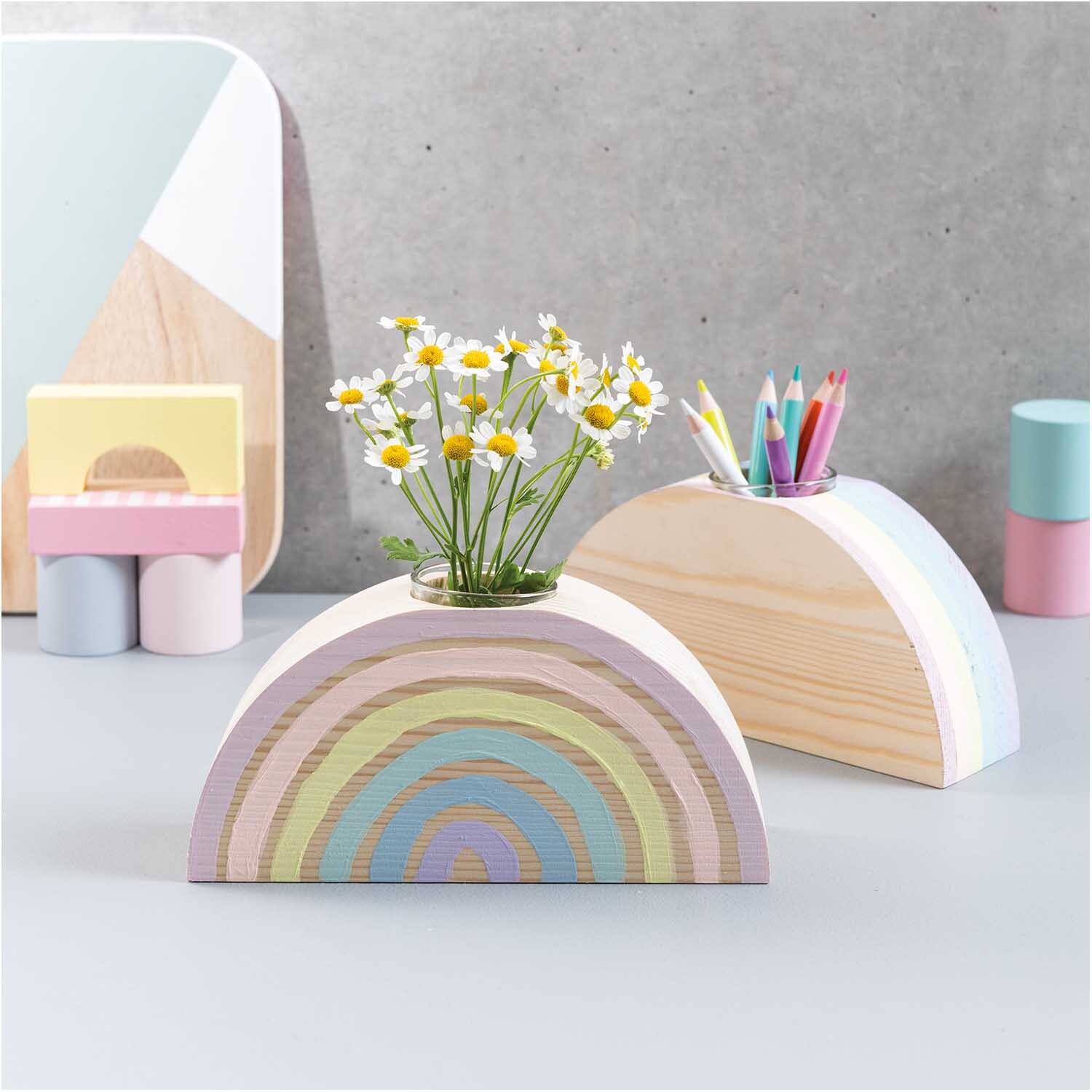 Holz-Regenbogen mit großer Glasvase, dekoriert mit Blumen, daneben bunte Stifte in einem ähnlichen Halter.