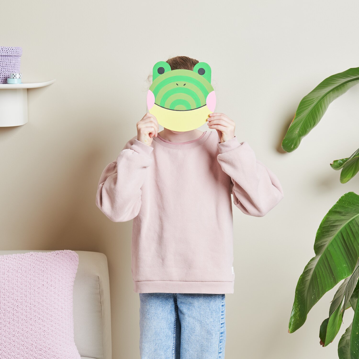 Malen nach Zahlen Frosch: Kind hält bunte Froschmaske vor Gesicht, trägt rosa Pullover und Jeans.