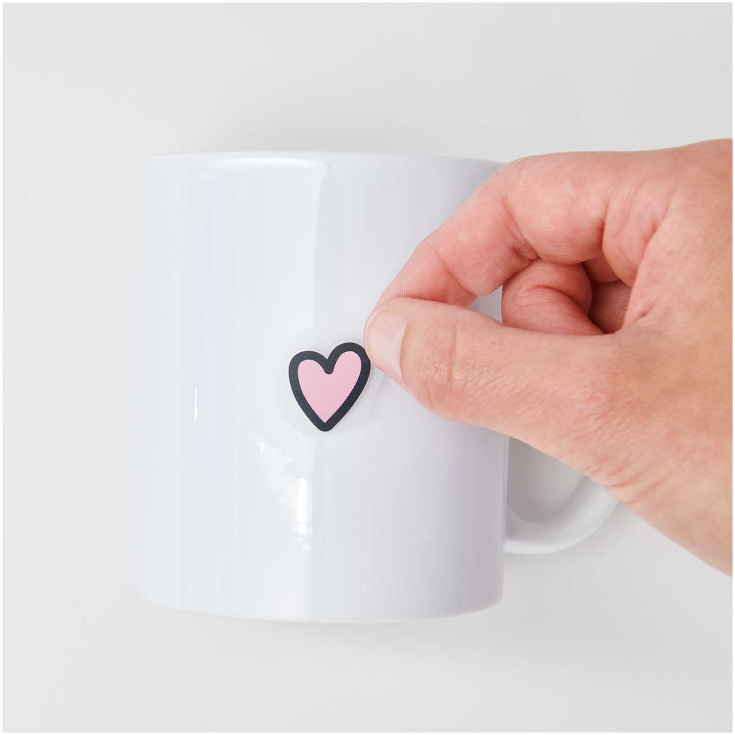 Transfermotiv Just Married: Eine Hand klebt ein rosa Herz auf eine weiße Tasse. Marke: Rico Design.