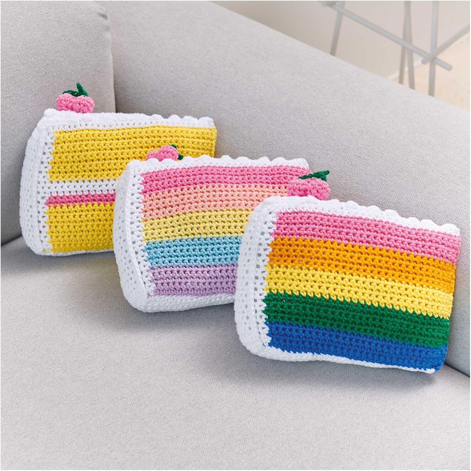 Creative Cotton aran: Drei gehäkelte Täschchen in Regenbogenfarben auf einem grauen Sofa. Ideal für kreative Projekte.
