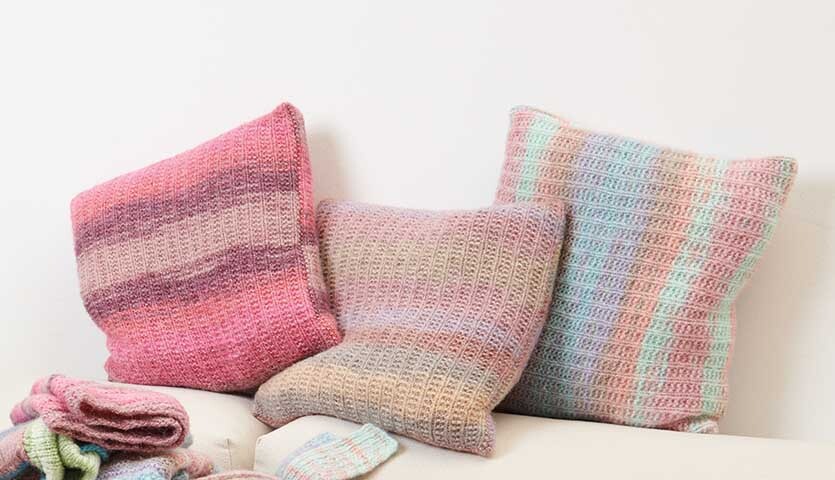 Strickanleitung Kissen aus Creative Painted Power: Drei gestrickte Kissen in Pastelltönen auf einem Sofa.