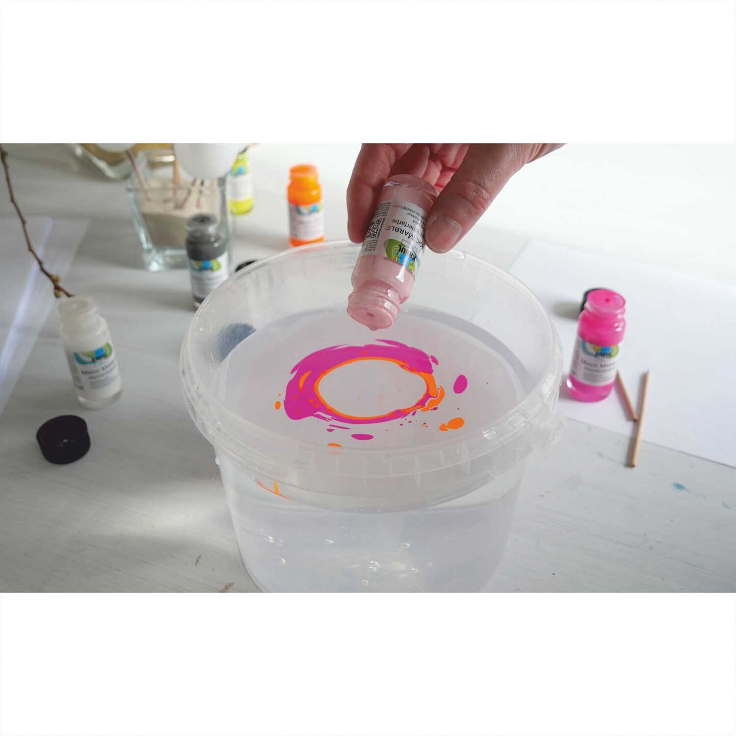Magic Marble Marmorierfarben Set Love Neon 6x20ml: Eine Hand tropft Farbe in Wasser, um kreative Muster zu erzeugen. Mehrfarbige Flaschen im Hintergrund.
