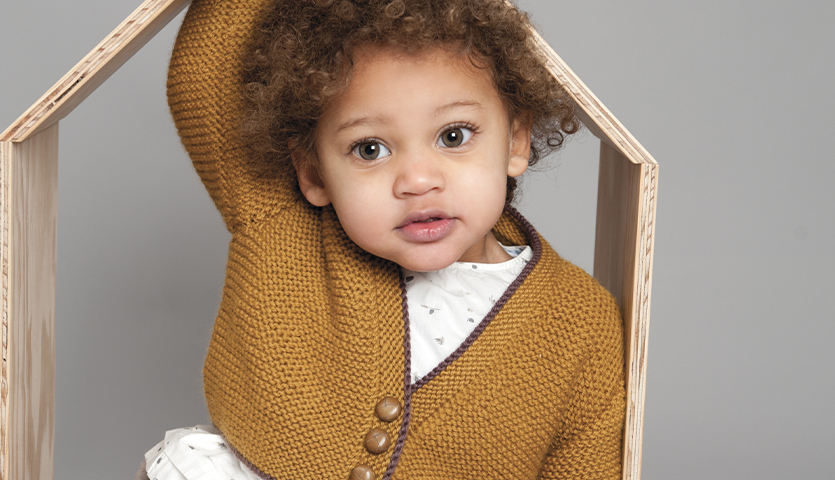 Strickanleitung Babybolero aus Essentials Cashlana: Kind trägt senfgelben Bolero vor grauem Hintergrund.