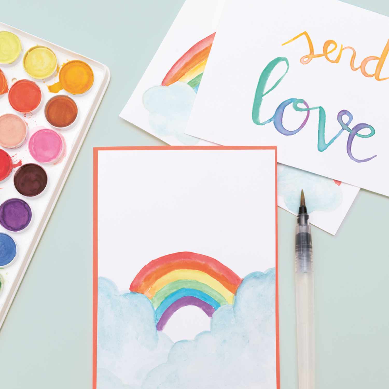 Anleitung kunterbunte Postkarten mit Aquarellfarben gestalten: Bunte Postkarten mit Regenbogenmotiv und Aquarellfarben-Set auf einem Tisch.