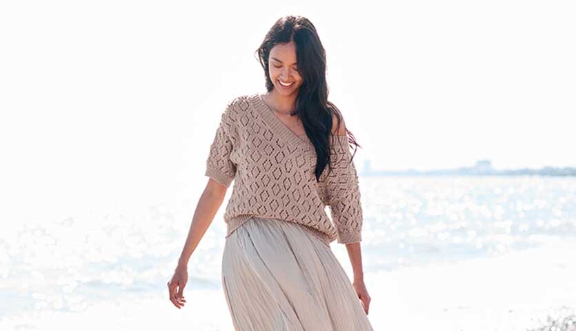 Strickanleitung Shirt aus Creative Silky Touch dk: Frau in beige gestricktem Oberteil am Strand.
