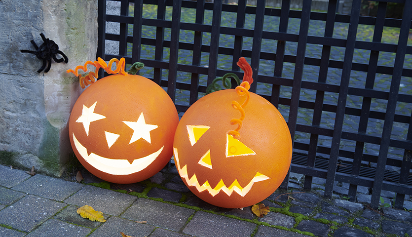 Bastelanleitung Halloweenkürbis aus Polystyrolkugel: Zwei leuchtende Kürbisse mit geschnitzten Gesichtern und Dekorationen vor einem Gitter.