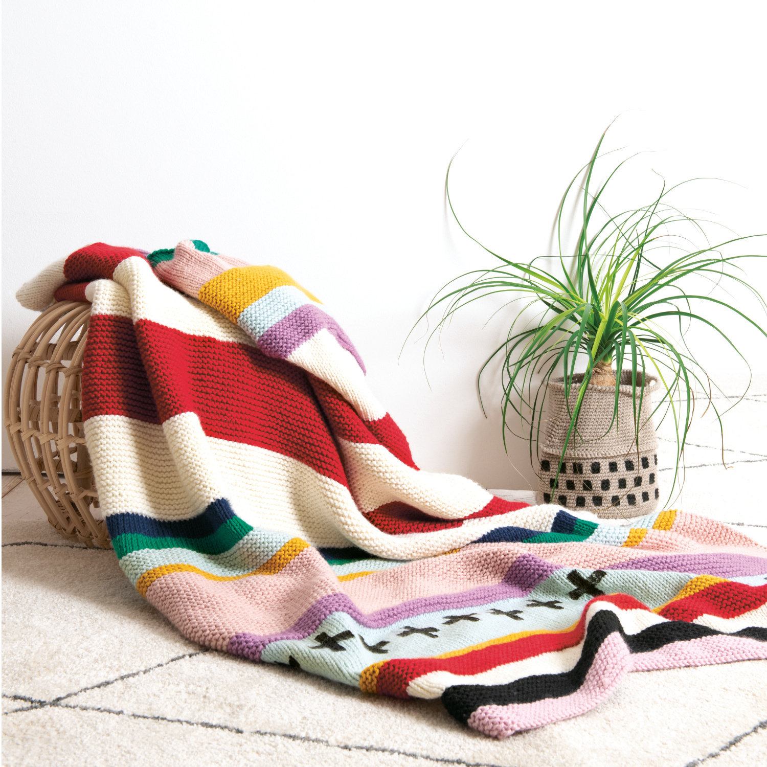 Strickanleitung Decke aus Essentials Mega Wool chunky: Bunte gestreifte Decke auf Korb, daneben grüne Pflanze auf hellem Teppich.