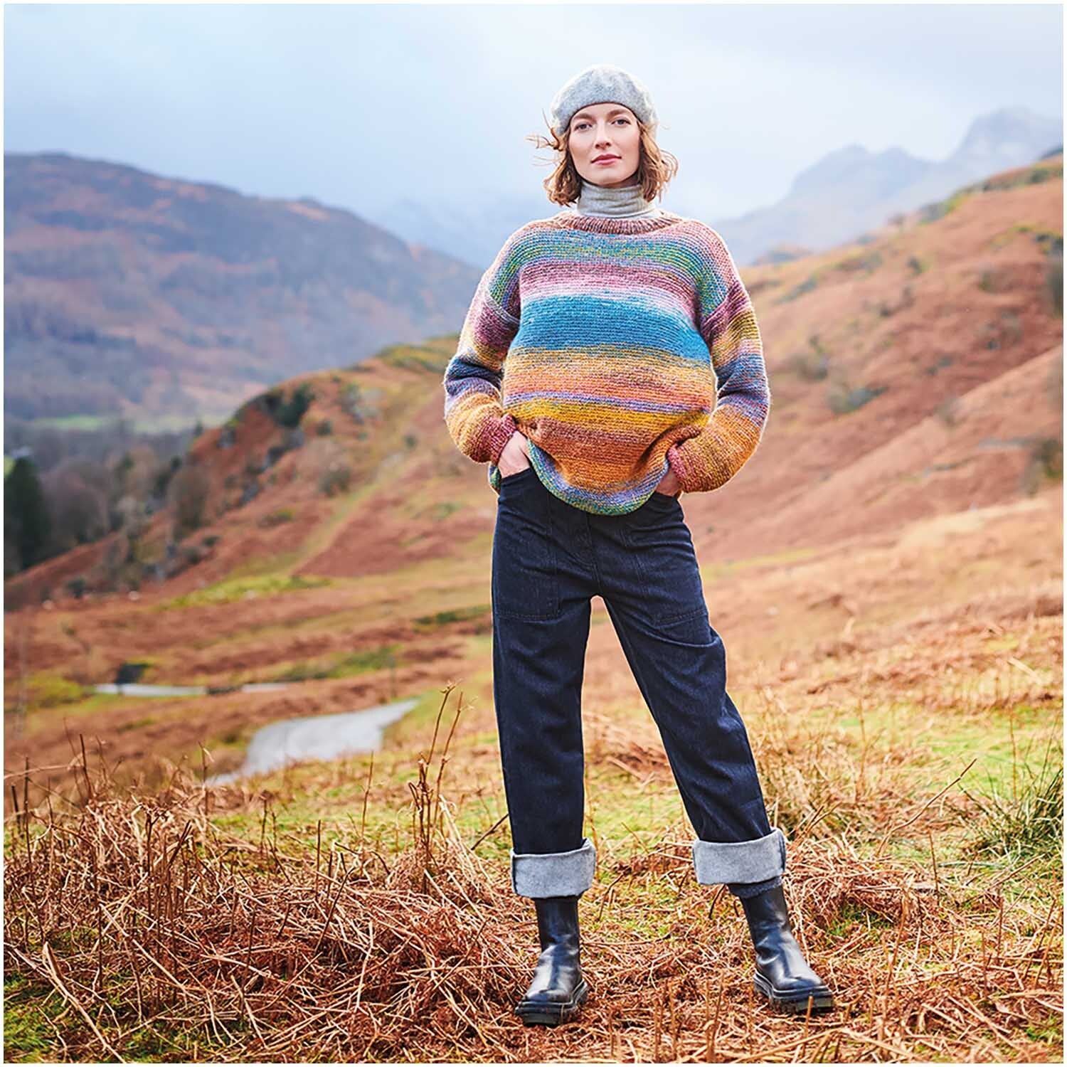 Strickset Pullover Modell 15: Bunter Pullover in Landschaft, getragen von Frau mit Mütze und Jeans.