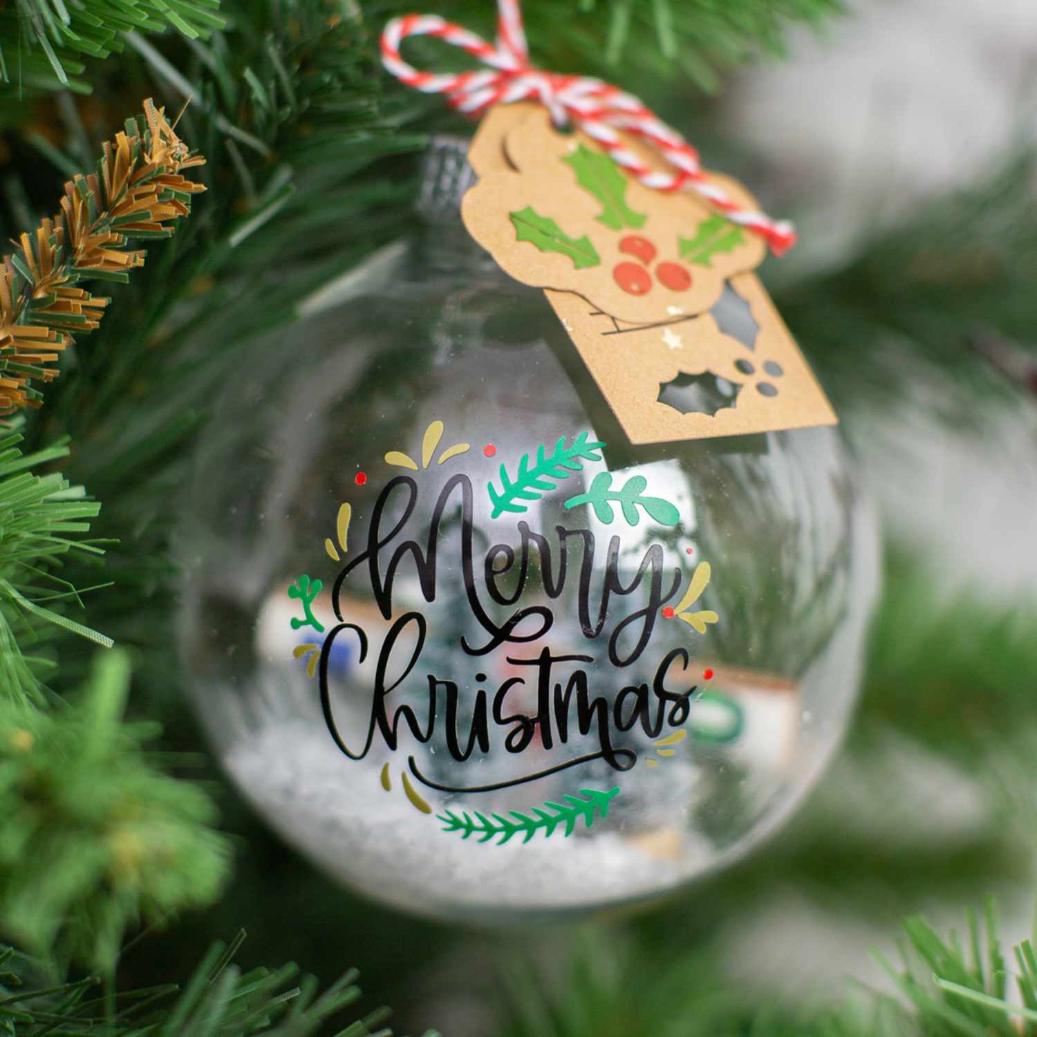 Bastelanleitung Weihnachtskugeln beplotten: Eine durchsichtige Kugel mit "Merry Christmas"-Schriftzug hängt an einem Tannenzweig.