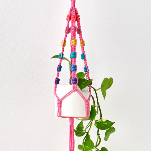 Weißer Blumentopf mit Pflanze hängt in einer pinken Blumenampel mit bunten Perlen. Fotosetting weiße Wand.