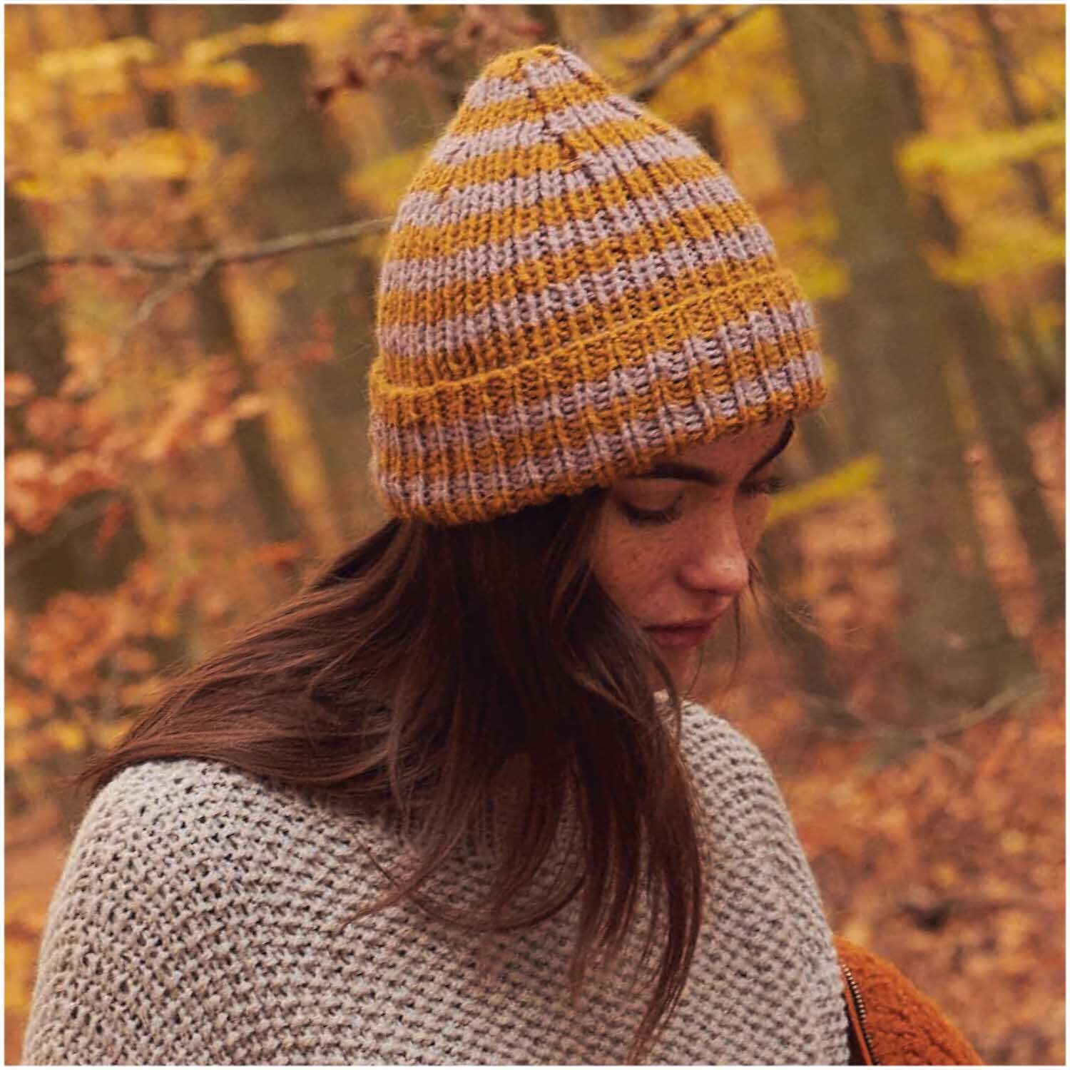 Essentials Alpaca Twist Chunky: Frau im Wald mit gestreifter Mütze aus Rico Design Wolle, Herbstfarben.