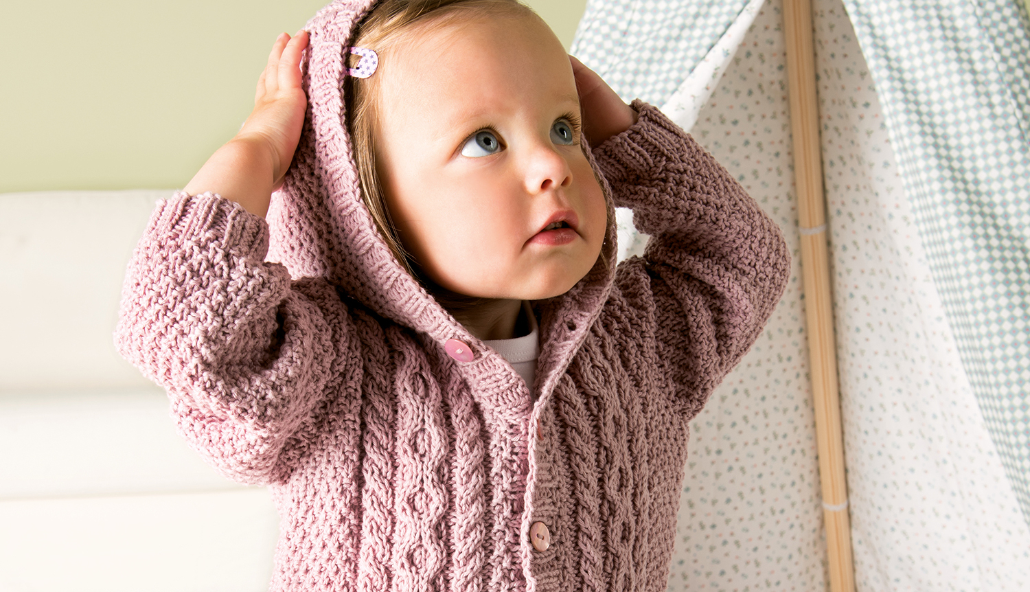 Strickanleitung Rico Baby 19 - Zopfjacke: Baby in rosa Zopfjacke mit Kapuze, hält die Kapuze fest.