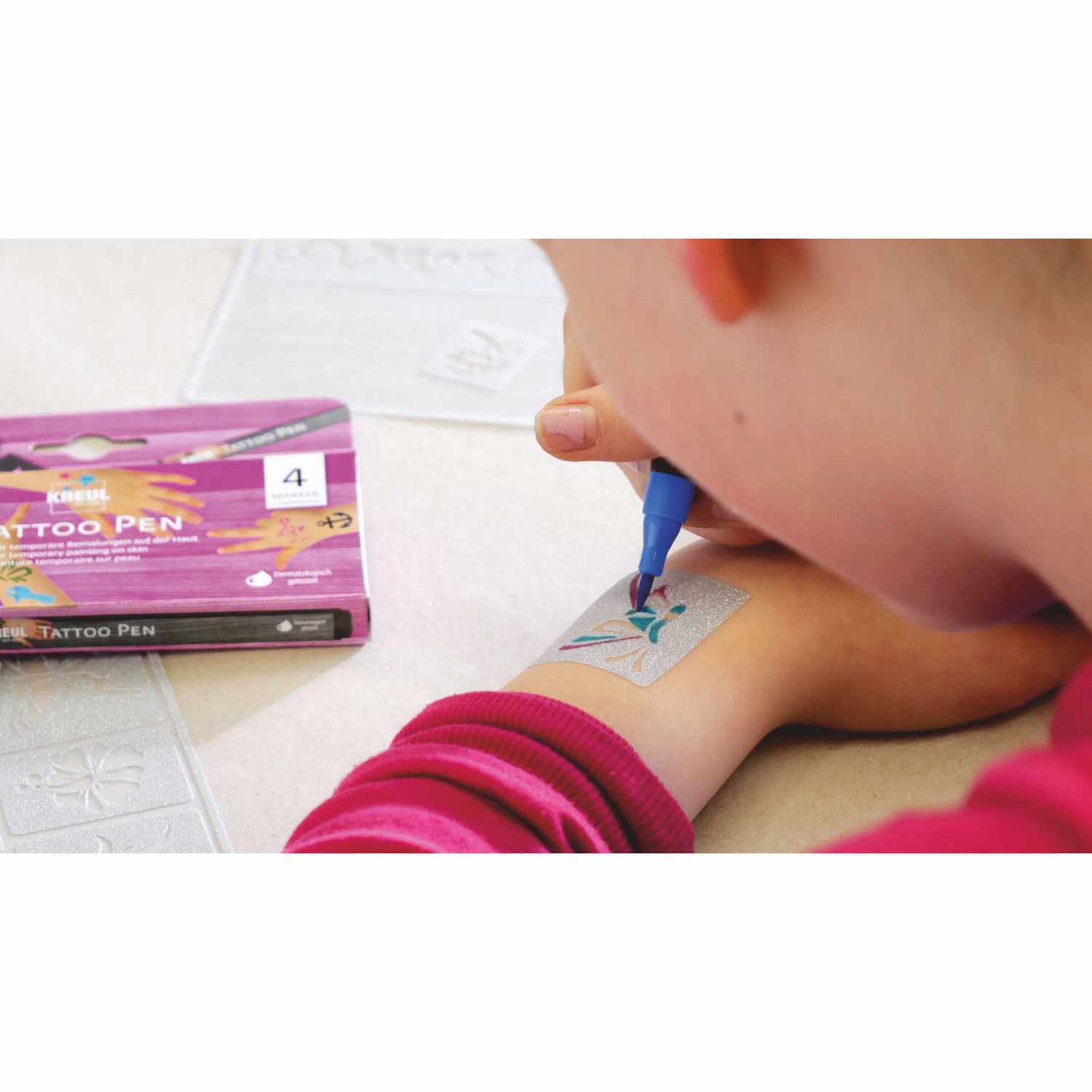 Tattoo Pen 4er-Set Anker, Sterne und Schmetterling: Kind malt mit blauem Stift auf Arm, KREUL-Set im Hintergrund.