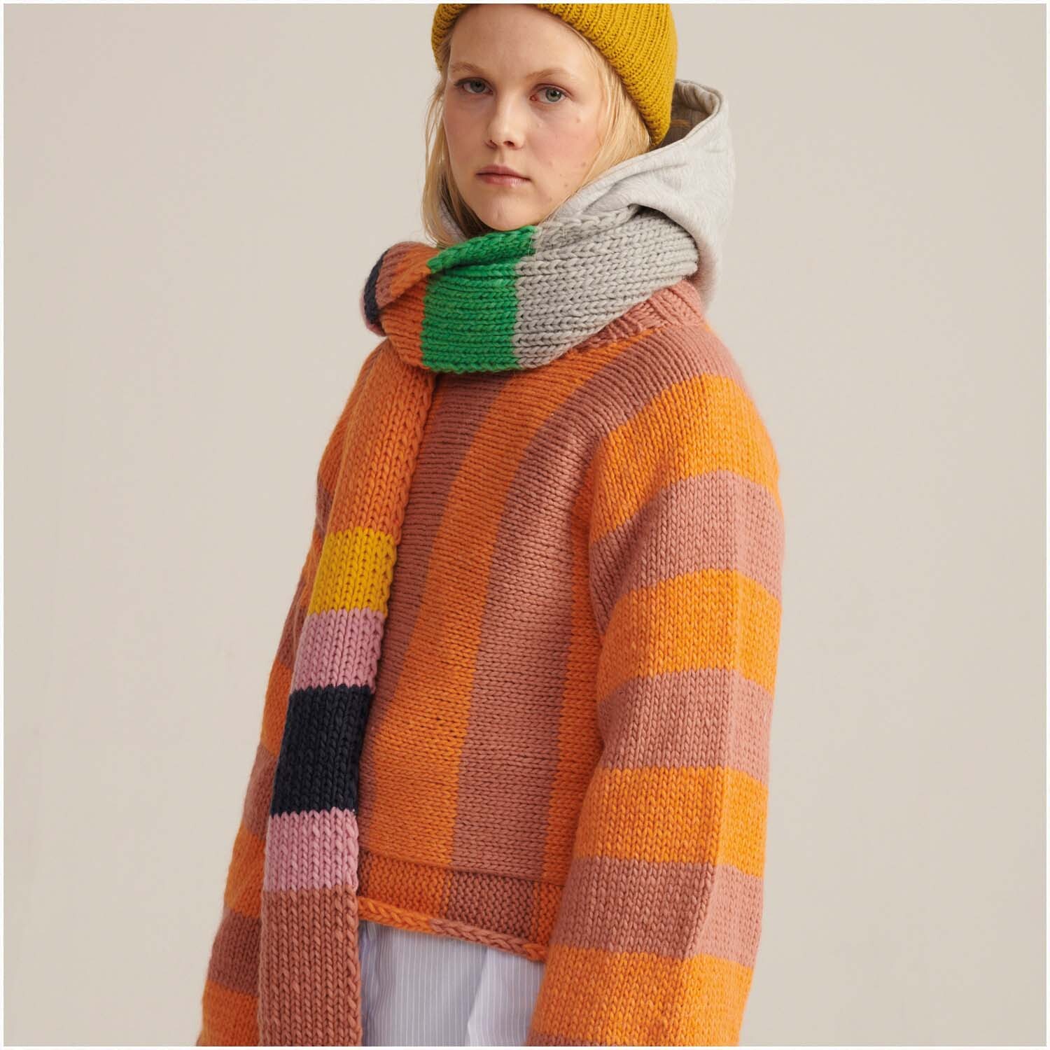 Essentials Super Chunky Pullover in Orange und Rosa von Rico Design. Frau trägt gestreiften Schal und gelbe Mütze.