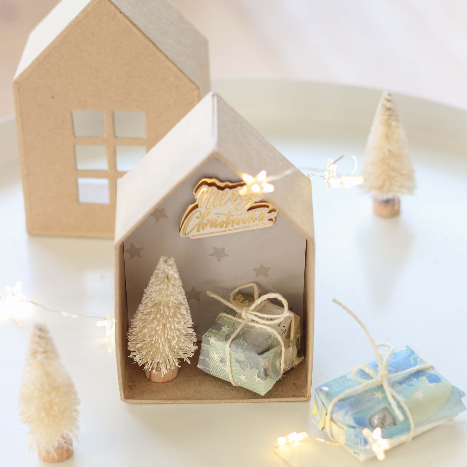 Bastelanleitung Geldgeschenk in Weihnachtskulisse: Miniaturhaus mit Geschenken und Tannenbäumen, festlich dekoriert.