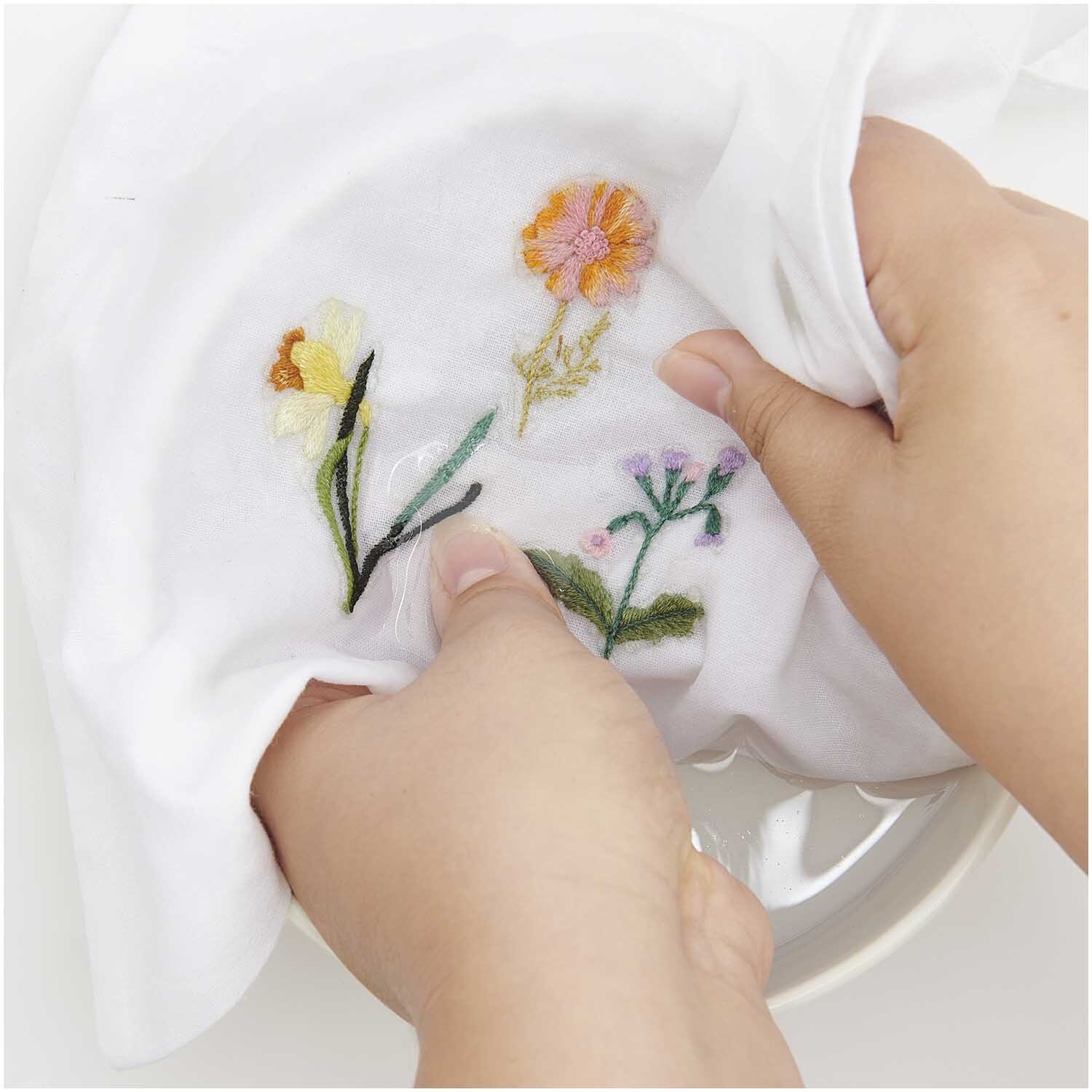 Stick and Stitch Stickpackung Frühlingsblumen zeigt bunte Blumenstickerei auf weißem Stoff. Hände halten das Tuch, ideal für kreative Projekte.