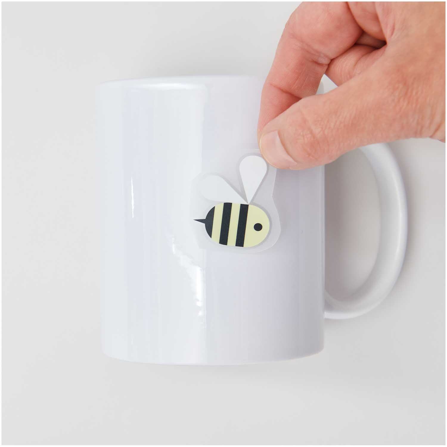 Transfermotiv Biene: Eine Hand klebt ein Bienenmotiv auf eine weiße Tasse. Marke: Rico Design.