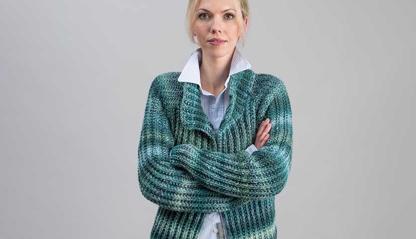 Strickanleitung Jacke aus Creative Ito Iro chunky: Frau trägt gestrickte, blau-grüne Jacke mit Schalkragen vor grauem Hintergrund.