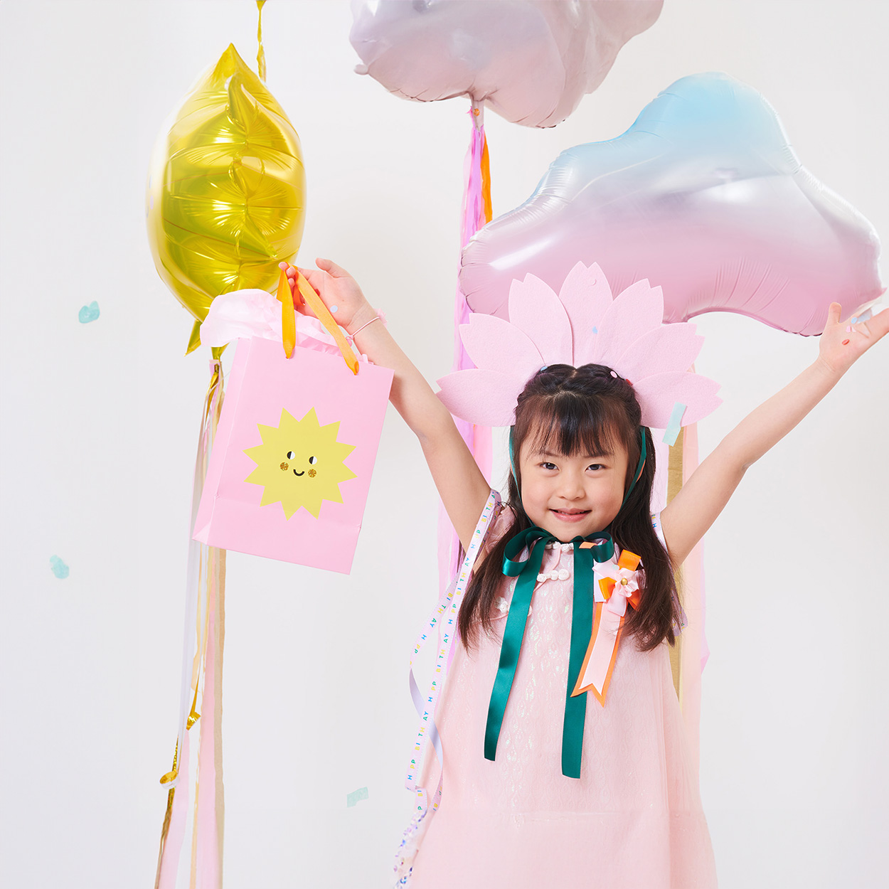 Ein Mädchen hebt die Hände in die Höhe und hält eine Geschenktüte in der Hand, außerdem trägt sie einen Kirschblüten Haarreif und hinter ihr sind Luftballons.