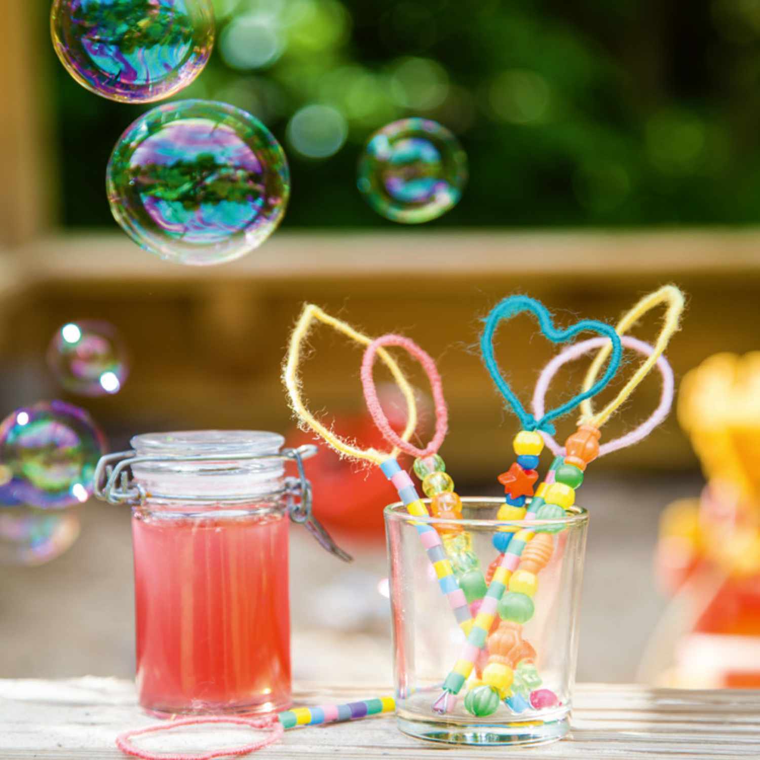 Bastelanleitung DIY-Seifenblasen: Bunte Seifenblasen schweben über einem Tisch mit Bastelutensilien und einem Glas mit farbigen Stäben.