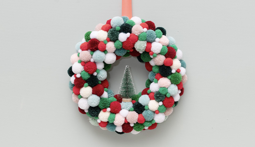 Bastelanleitung weihnachtlicher Pompon-Türkranz: Bunter Kranz aus Pompons mit kleinem Baum in der Mitte, ideal für festliche Dekoration.
