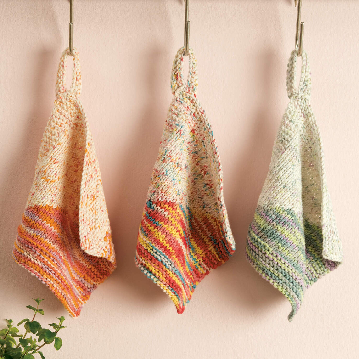 Strickanleitung Topflappen aus Creative Cotton Print: Drei bunte Topflappen hängen an Haken vor einer Wand.